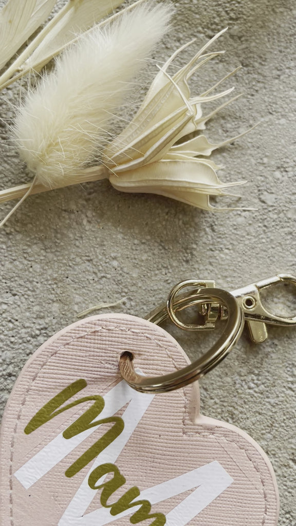 Personalisierter "Mama"-Herz-Schlüsselanhänger liegt neben Trockenblumen auf dem Boden
