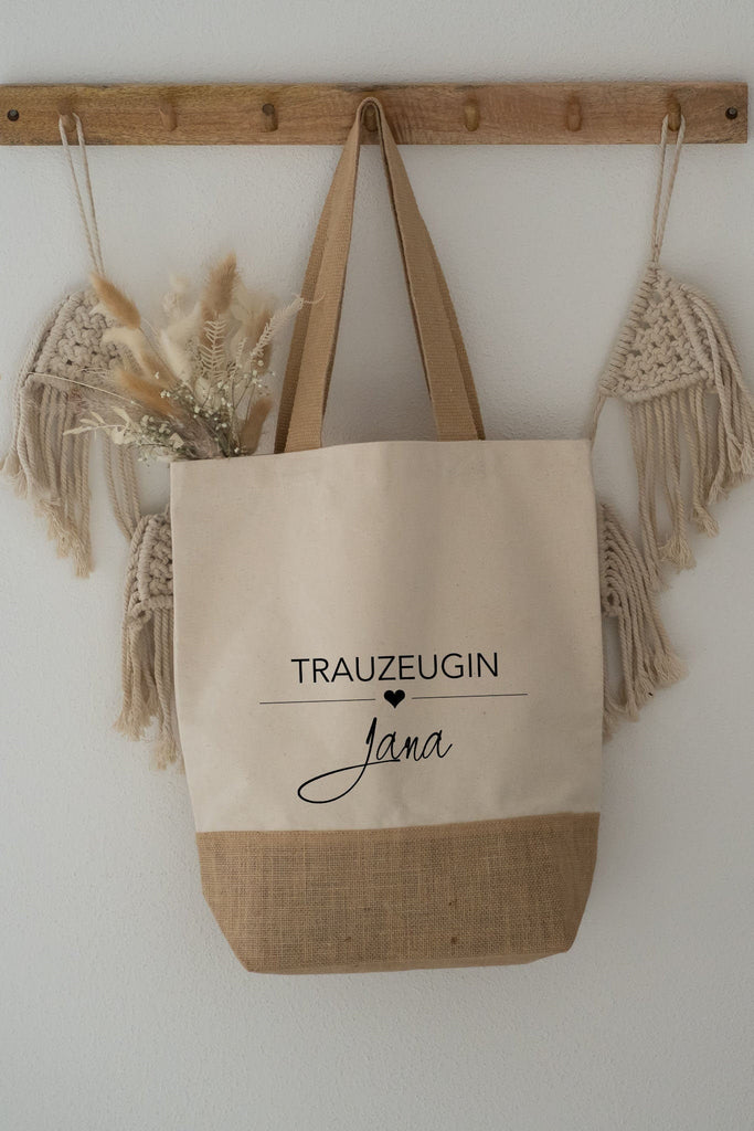 Personalisierte Jutetasche hängt an einem Holzhaken, Schilf und Trockenblumen gucken aus der Jutetasche heraus