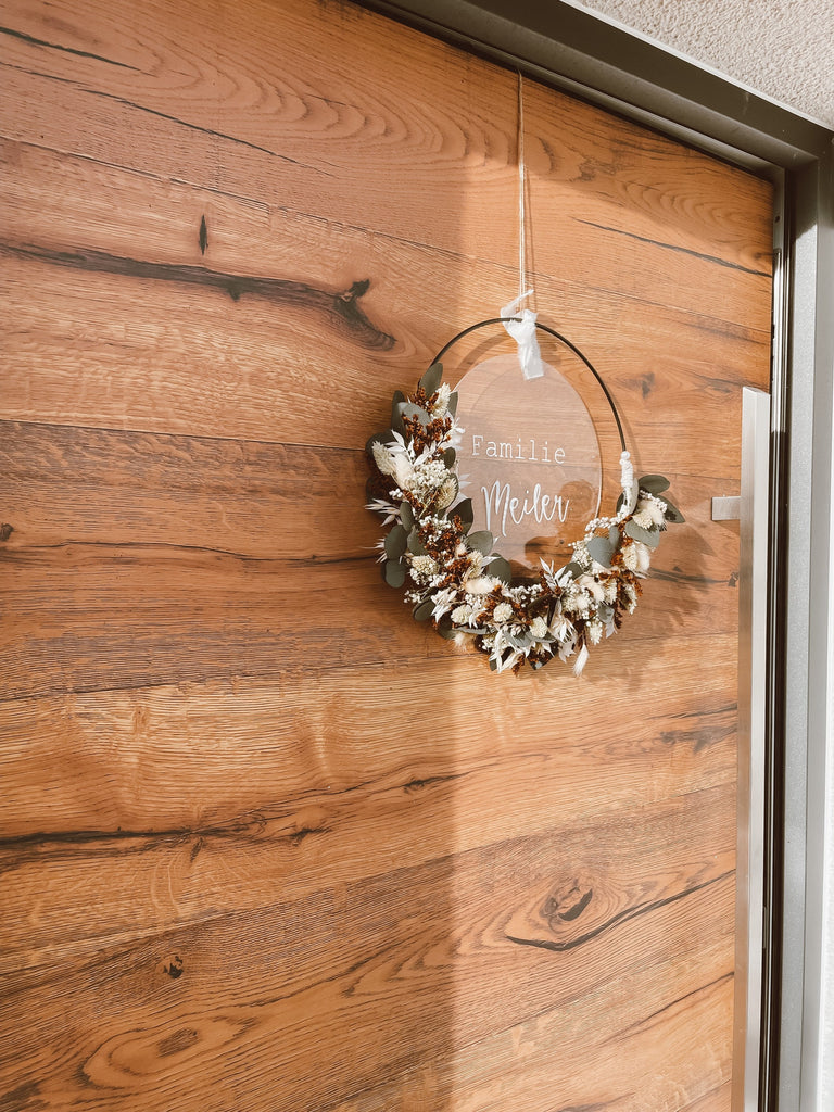 Trockenblumenkranz mit "Familie Meiler" personalisierter Acrylglasscheibe hängt an einer Tür