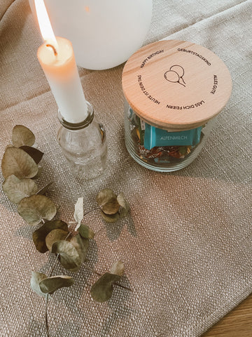 Tischdecke auf der ein Vorratsglas mit einem personalisiertem Holzdeckel, eine Kerze und Trockenblumen stehen