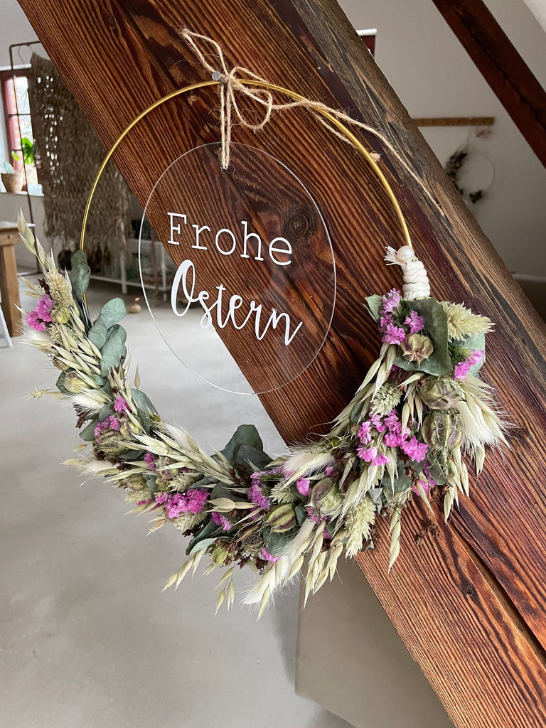 Trockenblumenkranz mit "Frohe Ostern" personalisierter Acrylglasscheibe  hängt an einem Holzbalken, Sofa und eine Palme stehen in dem Hintergrund