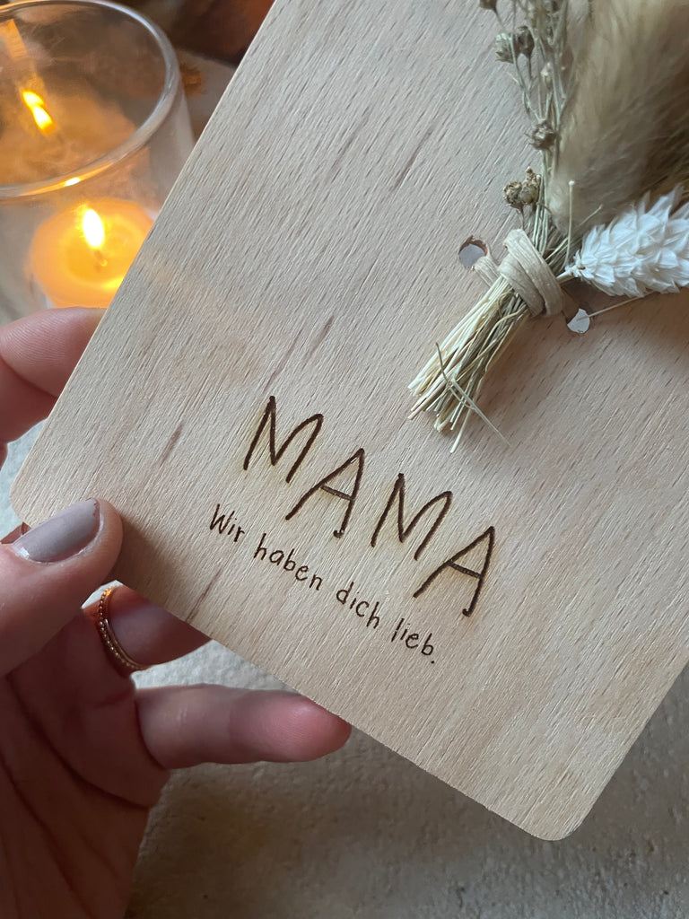 Nahaufnahme von einer Holzkarte mit der Gravierung "Mama Wir haben dich lieb", Brennende Kerze in einem Glas