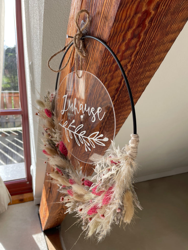 Trockenblumenkranz mit personalisierter "Zuhause" Acrylglasscheibe hängt an einem Holzbalken